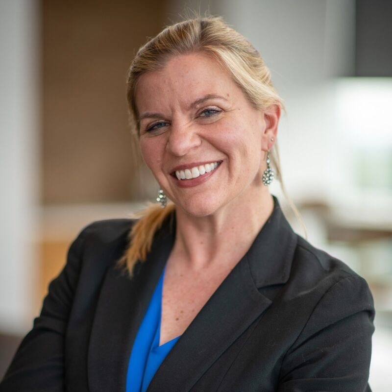 The image shows a woman with blonde hair pulled back, wearing a black blazer over a blue top. She is smiling at the camera. The background is blurred, suggesting an indoor setting. She appears to be in a professional environment.